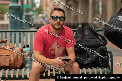 Downtown city sidewalk - July 2022: A man in sunglasses sits on a metal bench checking his phone, luggage and a backpack beside him on a city street.
