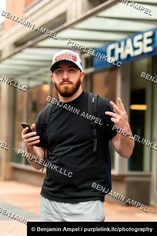 Downtown city sidewalk - July 2022: A man in a ball cap and backpack holds his phone and gestures while standing outside a bank branch storefront.