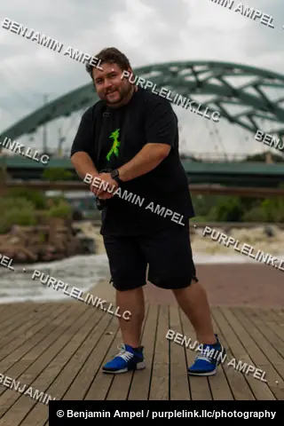 Riverside boardwalk - July 2022: A man poses on a wooden boardwalk beneath a large green steel arch bridge spanning the water behind him.