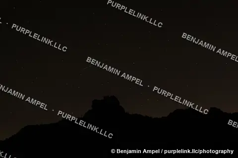 A bright meteor streaks diagonally across a star-filled night sky above the silhouette of a rugged desert mountain.