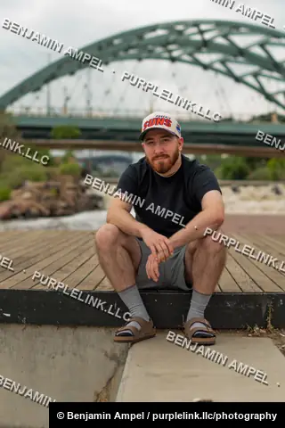 Riverside boardwalk - July 2022: A man in a sports cap and sandals sits on a concrete ledge near the water, a steel arch bridge rising behind him.