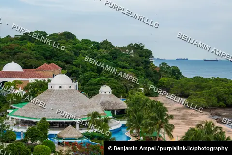 An aerial view of a tropical beach resort with thatched-roof pavilions and a pool, surrounded by jungle with cargo ships anchored offshore.
