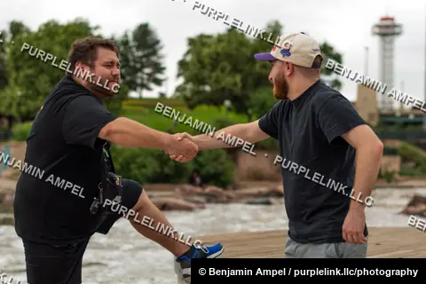 Riverside boardwalk - July 2022: Two men shake hands on a wooden boardwalk beside a river, a steel arch bridge and observation tower visible in the background.