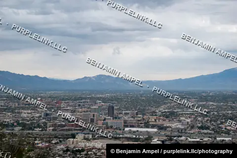A sprawling desert city skyline sits in a broad valley ringed by distant mountains, photographed from a high overlook under an overcast sky.