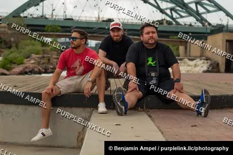Riverside boardwalk - July 2022: Three men sit together on a concrete ledge along a riverside boardwalk, a large steel arch bridge spanning the water behind them.