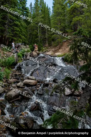 Rocky Mountain forest - July 2022: A waterfall cascades over moss-covered boulders in a pine forest, hikers resting on rocks near the top of the falls.