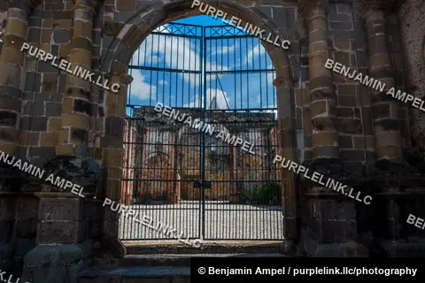 A locked wrought iron gate frames the view into the courtyard of crumbling stone colonial ruins beneath a bright blue sky.