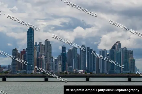 The dense modern skyline of Panama City rises above the water, viewed from across the bay under a cloudy sky.