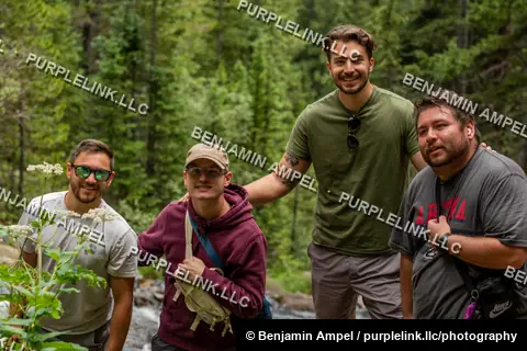 Rocky Mountain forest - July 2022: Four friends pose together smiling in a pine forest, a mountain stream visible behind them among wildflowers.