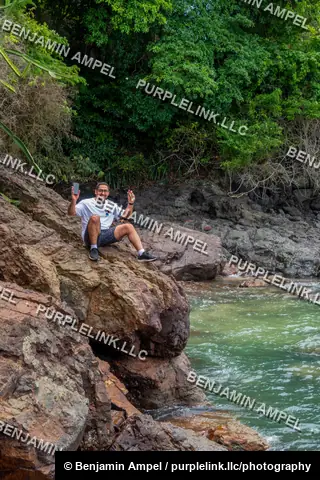 Panama - August 2023: A man in a white t-shirt sits on a rocky outcrop above a turquoise river, holding up his phone and giving a peace sign.