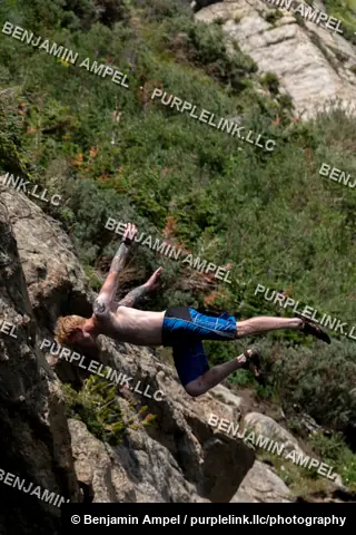 Rocky Mountain cliffs - July 2022: A man performs a backflip off a rocky cliff, arms outstretched and tattoos visible, greenery and granite rockface behind him.
