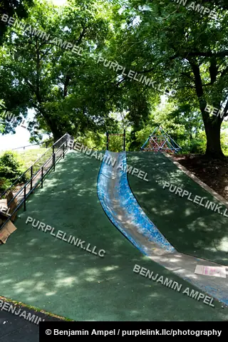 A blue plastic slide descends a grassy hill through dappled shade, surrounded by tall trees and simple playground equipment.