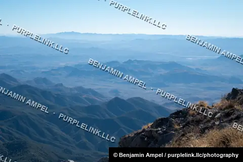 Layered mountain ridges fade into blue haze toward a distant valley, seen from a high rocky overlook.