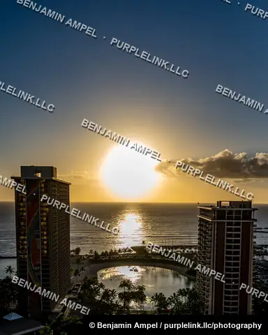 Waikiki, Honolulu, Hawaii - January 2024: The sun sets over the Pacific Ocean between illuminated high-rise towers, one decorated with a colorful mural, above a lagoon and palm trees.