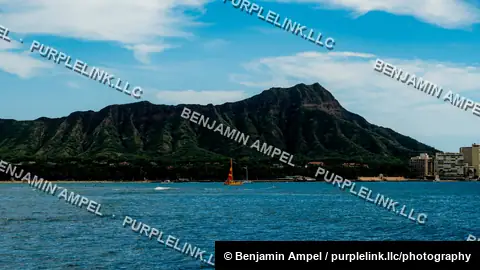 Diamond Head's rugged green slopes tower over the Waikiki shoreline, with a small sailboat crossing the blue water in the foreground.