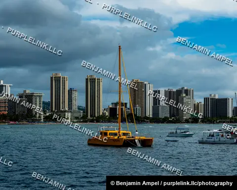 Waikiki, Hawaii - January 2024: A yellow sailing catamaran and tour boats float in front of the dense hotel and condo skyline of Waikiki Beach under a partly stormy sky.