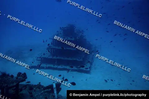 A tiered pyramid-shaped artificial reef structure sits on the sandy ocean floor, surrounded by schools of small fish, off the coast of Hawaii.