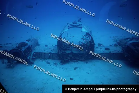 The corroded fuselage of a sunken vintage aircraft lies on a sandy seabed, encrusted with marine growth, off the coast of Hawaii.