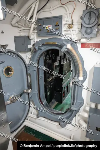 An open oval hatch on a submarine bulkhead reveals dense electrical equipment behind it, with an exit sign mounted on the wall above.
