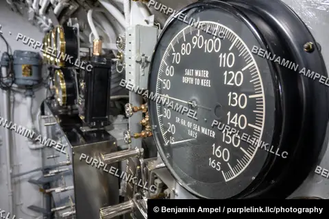 A close-up of a black and white depth gauge dial reading salt water depth to keel, mounted among other instruments inside a historic submarine.