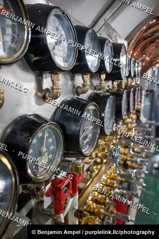 Dozens of round pressure gauges and brass valves line the wall of a submarine's control room, with red-handled valves in the foreground.