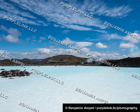 Iceland - June 2024: A milky turquoise geothermal spa lagoon stretches toward volcanic hills under a bright blue sky with scattered clouds.