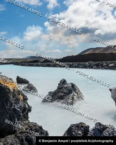 Iceland - June 2024: Volcanic rocks rise from milky turquoise geothermal water with steam drifting across the surface under a partly cloudy sky.