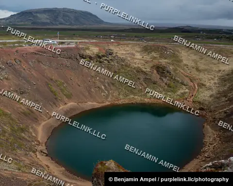Iceland - June 2024: A deep volcanic crater holds a still teal lake, its slopes striped with red volcanic soil and green vegetation, beneath a distant flat-topped mountain.