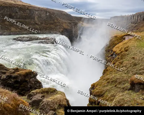 Iceland - June 2024: Gullfoss waterfall drops into a narrow, mist-filled canyon lined with moss-covered cliffs, with distant visitors on the far rim.