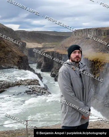 Iceland - June 2024: A young man in a gray jacket and beanie stands at a canyon overlook with Gullfoss waterfall's rushing river visible below.