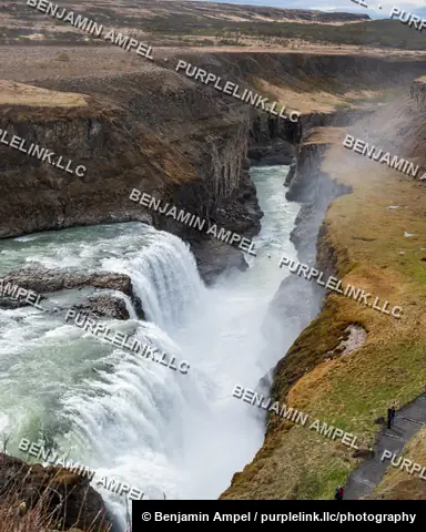 Iceland - June 2024: A vertical view down the misty canyon at Gullfoss waterfall shows small hikers walking a trail along the moss-covered rim.