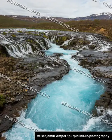 Iceland - June 2024: Bright blue glacial water cascades over dark volcanic rock ledges at Bruarfoss waterfall, surrounded by moss and distant snow-capped mountains.