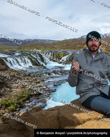 Iceland - June 2024: A young man in a beanie and jacket sits on a rock giving a thumbs up beside the bright blue Bruarfoss waterfall.