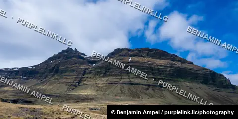 Iceland - June 2024: A rugged, layered mountain peak rises above green fields under a bright blue sky with scattered clouds.