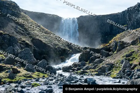 Snæfellsnes peninsula, Iceland: a waterfall drops through a green cliff into a rocky boulder-strewn stream amid drifting mist.