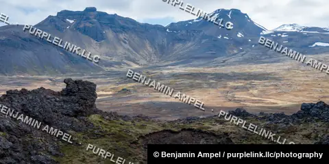 Wide valley of volcanic lava fields beneath snow-patched mountain ridges in the Icelandic interior.