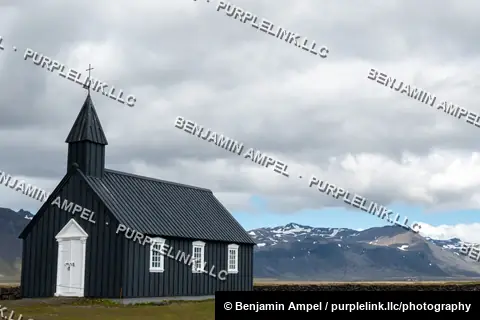 Búðakirkja, the black wooden church at Búðir, stands alone on green grassland with snow-dusted mountains behind, Iceland.