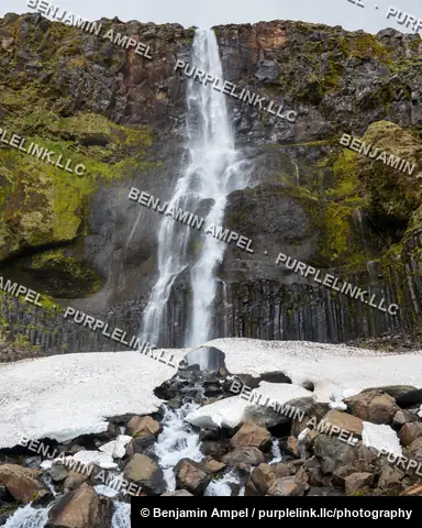 Bjarnarfoss waterfall cascades over dark basalt columns and a patch of snow, Snæfellsnes peninsula, Iceland.