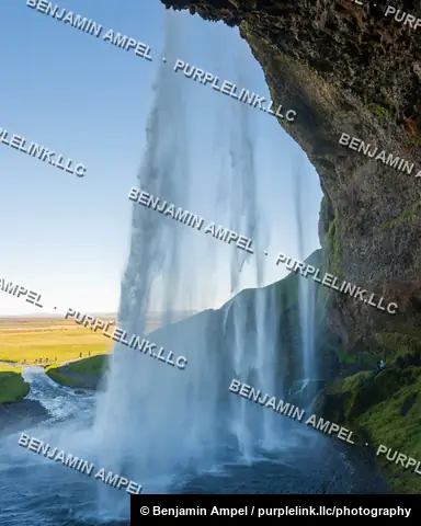 South Iceland - June 2024: sunlight streams through the curtain of Seljalandsfoss waterfall viewed from the cave behind, with visitors on the surrounding trail below.