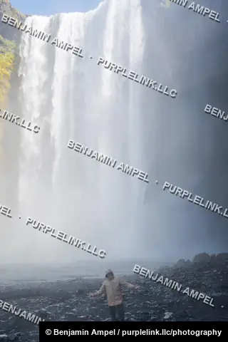 South Iceland - June 2024: a person in a rain jacket stands with arms outstretched at the base of Skógafoss waterfall amid rising mist.