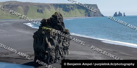 A dark basalt sea stack rises from a black sand beach with the Reynisdrangar rock spires visible along the south Iceland coastline.