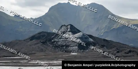 A striated, pyramid-shaped peak rises from a dark volcanic outwash plain beneath layered highland mountains, Iceland.