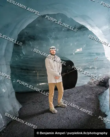 South Iceland - June 2024: a man in a helmet stands inside a glacier ice cave, surrounded by luminous blue and black striated ice walls.