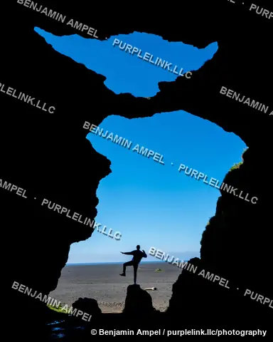 South Iceland - June 2024: a silhouetted figure strikes a pose beneath a rock cave arch opening onto a black sand beach and blue sky.
