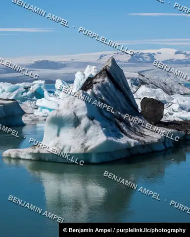 A dark, ash-streaked iceberg floats with its reflection in the calm waters of Jökulsárlón glacier lagoon, Iceland.