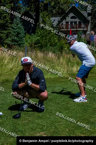Two men in golf attire stand on a fairway at a mountain golf course, one crouched near his club preparing to putt while the other swings.