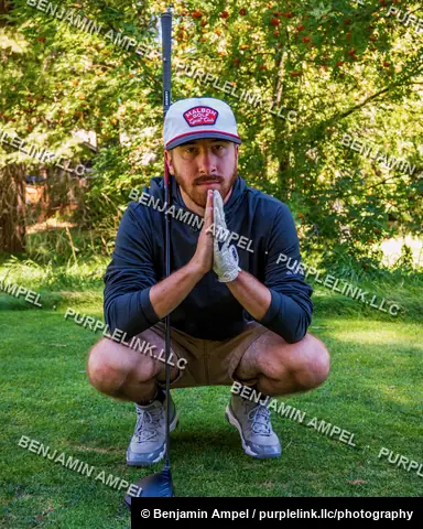 A man in golf attire crouches on a green lawn with hands clasped together in a playful pose, golf club resting beside him.