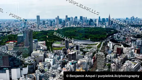 A wide aerial view over Tokyo shows the curved roof of Yoyogi National Stadium surrounded by green parkland with the dense city skyline beyond.