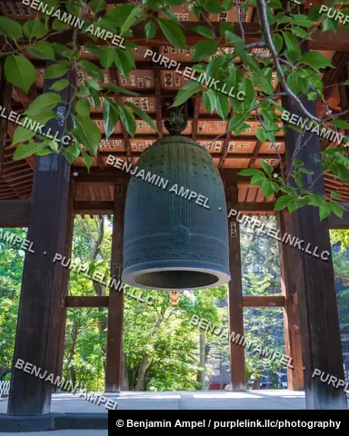 A large weathered bronze temple bell hangs inside a wooden pavilion, framed by green tree branches at a Buddhist temple.