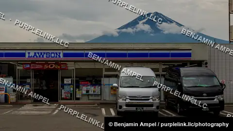 The snow capped peak of Mount Fuji looms above a Lawson convenience store parking lot with delivery vans parked outside.
